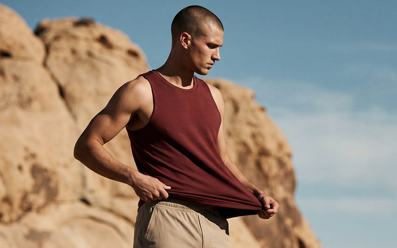 A man wearing men's active runner's apparel: a sleeveless burgundy tank top and khaki shorts stands outdoors in front of large tan rock formations under a blue sky, pulling the tank top slightly outward with both hands.